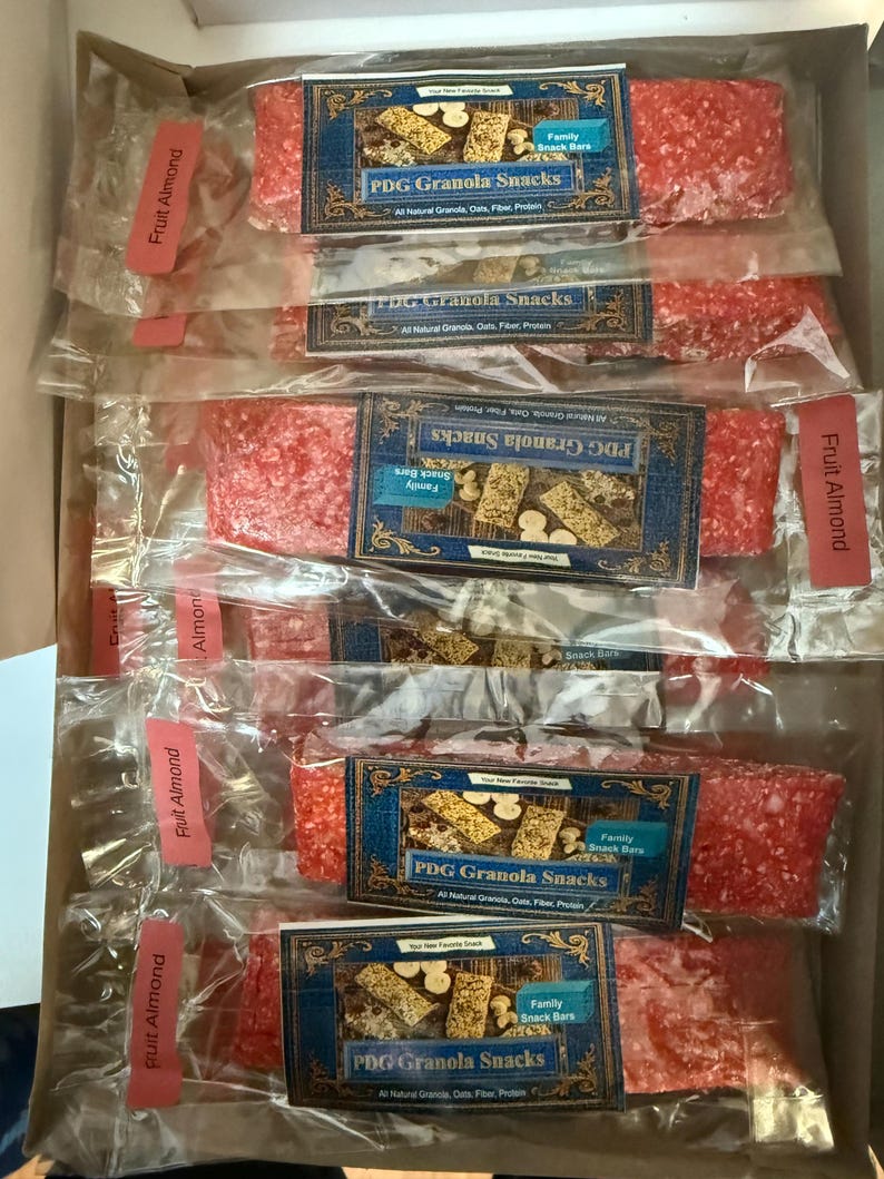 Może przedstawiać: Pudełko wypełnione pojedynczo zapakowanymi przekąskami PDG Granola, oznaczone jako "Fruit Almond". Każdy baton ma żywy czerwony kolor i jest zapakowany w przezroczysty plastik. Etykiety mają niebiesko-złoty wz&oacute;r z tekstem.
