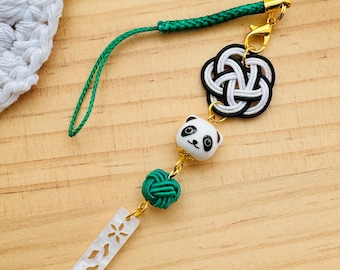 May include: A decorative charm featuring a panda bear, a green knot, and a clear rectangular pendant with floral cutouts. The charm is attached to a green cord and a gold-colored clasp. The background is a light brown wooden surface.