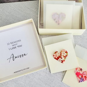 Könnte beinhalten: Eine quadratische cremefarbene Geschenkbox mit einem Stapel kleiner weißer Karten. Eine Karte trägt den Text 10 REASONS WHY I LOVE YOU Anissa. Weitere Karten zeigen einzelne gefaltete Papierherzen mit Blumenmustern in Rot-, Rosa- und Weißtönen.