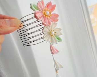 May include: A hair comb featuring two fabric flowers in light pink and white, each with a central pearl bead and yellow stamens. Small light green fabric leaves are attached to the flowers, and a dangling strand with two fabric petals and a white bead.