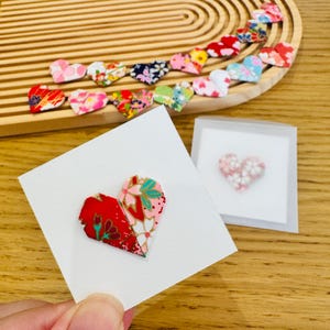 May include: A collection of small origami hearts crafted from patterned paper in various colours including red, pink, green, and blue. One heart is attached to a square white card, while another is inside a clear sleeve, and others are arranged.