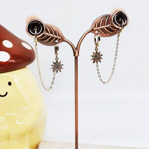 May include: A pair of gold star earrings with a chain dangle. The earrings are displayed on a brown metal stand.
