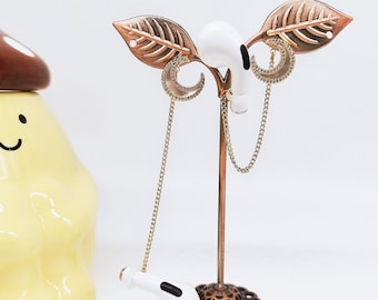 May include: A white wireless earbud with a gold chain and crescent moon charm hanging from a gold leaf stand.