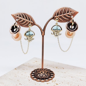 May include: A set of four earrings featuring a jellyfish design with blue and green accents. The earrings are displayed on a decorative stand with a leafy design.