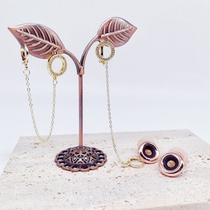 May include: A gold metal earring display stand with two pairs of earrings. The earrings are gold with clear stones. The stand is on a light brown textured surface.