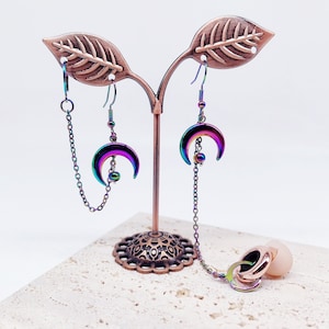 May include: A pair of iridescent rainbow crescent moon earrings with dangling chains. The earrings are displayed on a decorative stand with a leaf design.