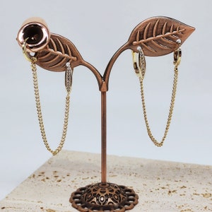 May include: A pair of gold hoop earrings with a small diamond accent hanging from a gold chain on a copper leaf-shaped stand.