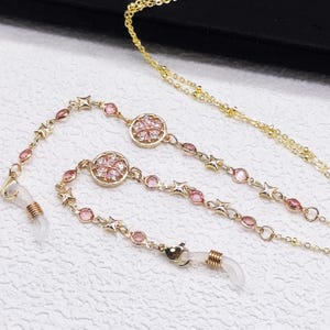 Op de afbeelding: Een goudkleurige brillenketting met delicate stervormige schakels, kleine ronde roze kristallen en cirkelvormige bedels met bloemmotief. De ketting is voorzien van doorzichtige siliconen lussen met goudkleurige metalen spiralen aan de uiteinden.