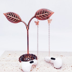 May include: A pair of gold butterfly earrings hanging from a decorative metal stand with a leaf design. The earrings are on a chain and have a delicate butterfly charm.