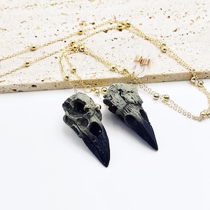 May include: A gold chain necklace with two black and green bird skull pendants. The chain has small gold beads.