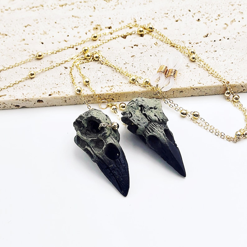 May include: A gold chain necklace with two black and green bird skull pendants. The chain has small gold beads.