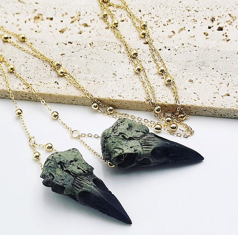 May include: Two gold chain necklaces with green and black bird skull pendants. The chains are made of small gold beads.