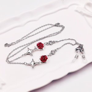 May include: A silver chain with two red rose charms and two silver star charms. The chain has clear crystal accents and two clear plastic eyeglass holders.