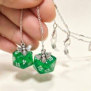 May include: A pair of eyeglass chains featuring silver-toned bee charms attached to translucent green twenty-sided dice with white numbers. The chains include silver metal links and clear silicone loops at the ends for securing to eyewear frames.