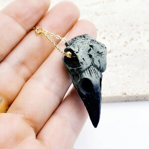 May include: A black and green crow skull pendant necklace with a gold chain. The skull is detailed and realistic, with a textured surface.