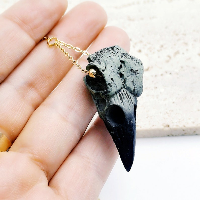 May include: A black and green crow skull pendant necklace with a gold chain. The skull is detailed and realistic, with a textured surface.