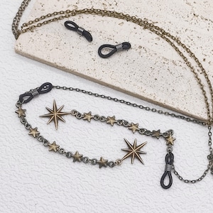 May include: A bronze-toned metal eyeglass chain featuring a series of small star charms and two larger eight-pointed star pendants. The chain includes black rubber loops at each end for securing to eyewear frames, with a delicate link design.