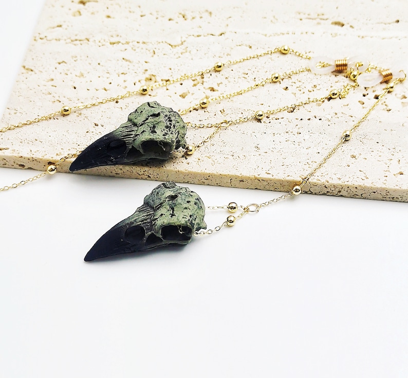 May include: A pair of long, gold chain necklaces with green and black bird skull pendants. The chains have small gold beads.