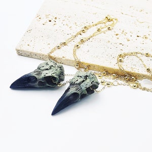 May include: A gold chain necklace with two green and black bird skull pendants. The chain has small gold beads.