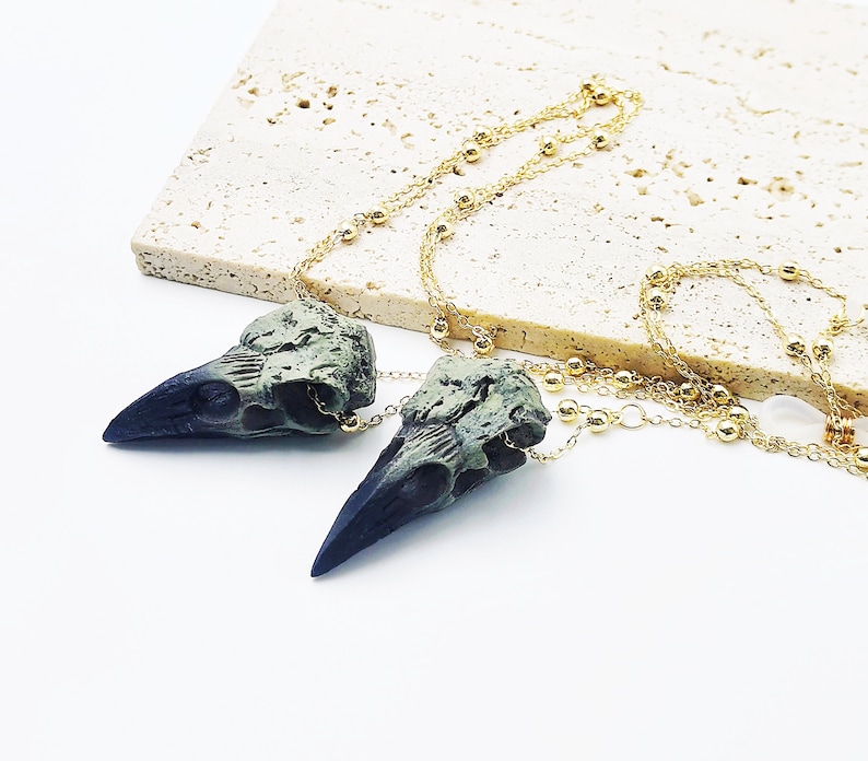 May include: A gold chain necklace with two green and black bird skull pendants. The chain has small gold beads.