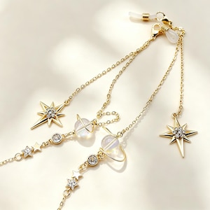 Puede incluir: Una cadena para gafas en tono dorado con dijes celestiales que incluyen estrellas de múltiples puntas con cristales transparentes, pequeñas estrellas recortadas y cuentas esféricas rodeadas por anillos, con bucles de silicona blanca.