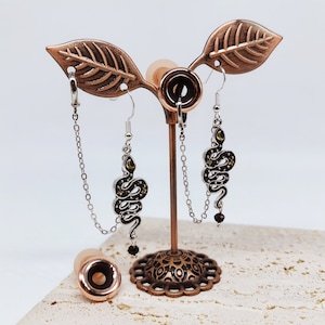 May include: A pair of silver snake earrings with gold accents hanging from a copper leaf earring display stand. The earrings are on silver chains and feature a black snake with gold stars.