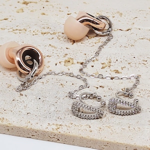 May include: Earrings with a chain and earbud-shaped accents. The earrings have a silver chain and are connected to two ear cuffs with a rectangular design, encrusted with small, clear stones. The earbud-shaped accents are a light pink color with rose gold accents.
