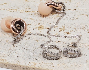 May include: Earrings with a chain and earbud-shaped accents. The earrings have a silver chain and are connected to two ear cuffs with a rectangular design, encrusted with small, clear stones. The earbud-shaped accents are a light pink color with rose gold accents.