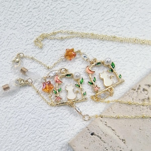 Op de afbeelding: Goudkleurige brillenketting met twee witte kattenhangers op een tak, geaccentueerd met oranje sterren, rode halve manen en groene bladeren. De ketting bevat transparante, met kralen bezette brilhouders en een delicaat kettingontwerp. Een charmant accessoire.