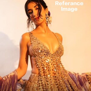 May include: A gold-colored, intricately beaded gown with a plunging neckline and open sides. The dress features a flowing skirt and is paired with matching gold jewelry. The image has a warm, golden hue.