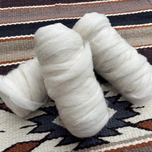 Op de afbeelding: Drie rollen witte wolvlies liggen op een patroontextiel. De wol is strak gewikkeld en heeft een zacht, getextureerd oppervlak. Het textiel heeft een geometrisch ontwerp in bruin, zwart en crème.