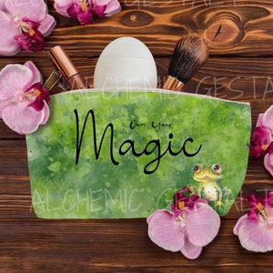 May include: A green zippered pouch with a watercolor design and the text "Own Your Magic". A frog illustration is on the pouch. The pouch is open and contains makeup brushes, a bottle of lotion, and a tube of lipstick. Pink orchid flowers surround the pouch.