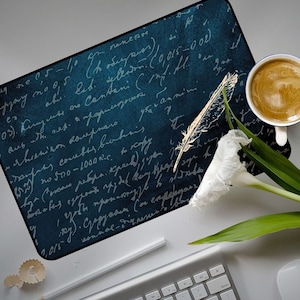 Puede incluir: Un mousepad azul oscuro con escritura manuscrita y un diseño de pluma dorada. También se ven una flor blanca, una taza de café y un teclado. El mousepad tiene una estética vintage y antigua.