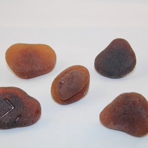 May include: Five smooth, brown sea glass stones, some with a darker brown or black hue. The stones are various shapes and sizes, with one featuring a carved symbol.
