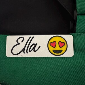 May include: White name tag with black text that reads "Ella" and a yellow smiley face with red hearts for eyes.