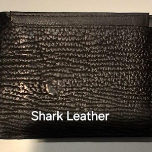 May include: A black shark leather wallet with a textured finish. The bi-fold wallet features a card slot. The text "Shark Leather" is printed in white on the front. The wallet is made of shark leather.