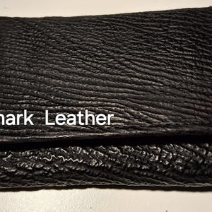 Peut inclure: Un portefeuille en cuir de requin noir avec une surface texturée. Les mots "Shark Leather" sont imprimés en blanc sur le devant. Le portefeuille est fermé et semble neuf.