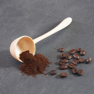 Può includere: Un cucchiaio di legno chiaro per caffè con manico, accanto a un mucchio di caffè macinato e diversi chicchi di caffè interi. I chicchi di caffè sono marrone scuro e il caffè macinato è marrone chiaro.