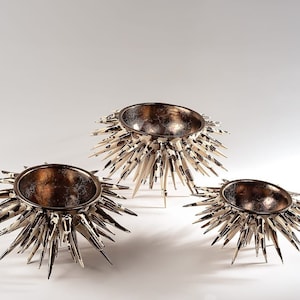 Può includere: Tre ciotole decorative con un interno in bronzo metallizzato e un esterno appuntito. Ogni ciotola presenta numerosi elementi sporgenti e acuminati con un motivo maculato color crema e nero, che ricordano i ricci di mare, in varie dimensioni.
