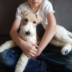 Pode incluir: Uma figura de cão feita em feltro de agulha com uma pelagem branca, castanha e preta. O cão tem uma aparência texturizada semelhante à lã, olhos escuros e um nariz preto. É moldado para se assemelhar a um terrier com orelhas dobradas.