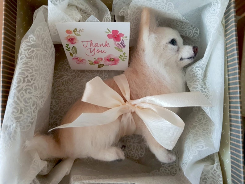 May include: A handcrafted dog figurine in light colours, presented in a decorative box with a cream-coloured bow. A small card with floral details and the words "Thank You" is included. The dog has a fluffy texture.
