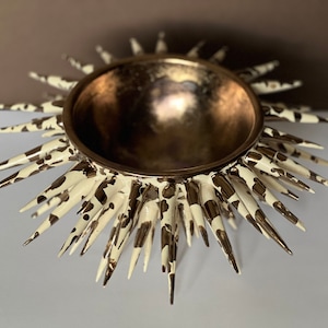 May include: Decorative sunburst bowl with a metallic gold interior and a cream and brown spotted exterior. The bowl has a circular shape with pointed rays extending outwards, creating a starburst effect. This decorative piece is ideal for home decor.