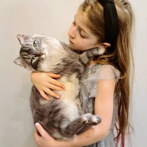 May include: A realistic, life-sized needle felted cat sculpture featuring long, soft-textured grey and white fur, pointed ears, and green eyes. The handcrafted feline figure is designed with a lifelike posture, capturing the appearance of a fluffy cat.