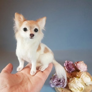 May include: A handcrafted miniature Chihuahua dog figurine. The dog has a white and tan coat, with a long, fluffy tail. The dog is sitting upright, with its head facing forward. The figurine is placed on a person's hand.