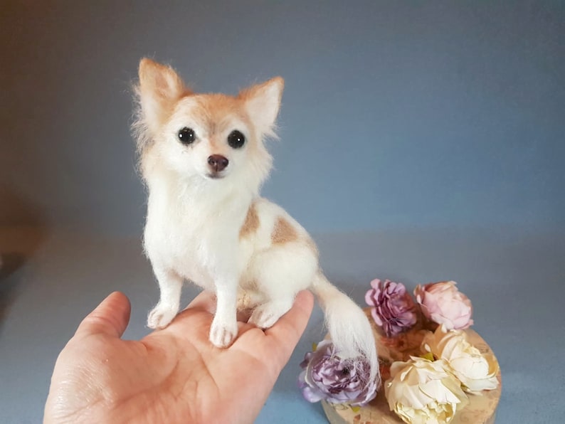 May include: A handcrafted miniature Chihuahua dog figurine. The dog has a white and tan coat, with a long, fluffy tail. The dog is sitting upright, with its head facing forward. The figurine is placed on a person's hand.