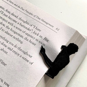 May include: A black bookmark in the shape of a person pointing, inserted into a book. The book is open to page 61, with text visible. The bookmark is made of a solid, opaque material.