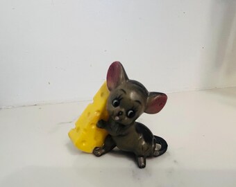 May include: A small figurine of a cartoon mouse in a crouching position, holding a large yellow cheese wedge. The mouse is gray with pink ears and a black tail. The cheese has holes.
