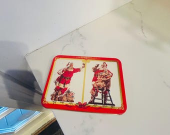 May include: A rectangular metal tin featuring a red border and two illustrations of Santa Claus. One Santa stands with a sack of toys, while the other sits on a stool holding a sack. The design includes gold accents and decorative ribbon motifs.