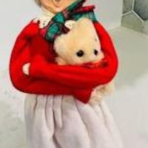 May include: A vintage-style doll wearing a red dress, white apron, and a white bonnet. She is holding a white teddy bear in her arms.