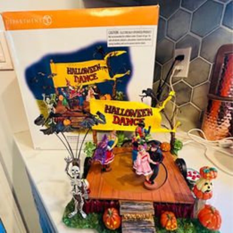 Op de afbeelding: Een muzikaal dorpssc&egrave;ne met Halloween-thema met een draaiend podium met een skelet, een heks en een dansend stel. Het podium is versierd met pompoenen, vleermuizen en een banner met de tekst "Halloween Dance".